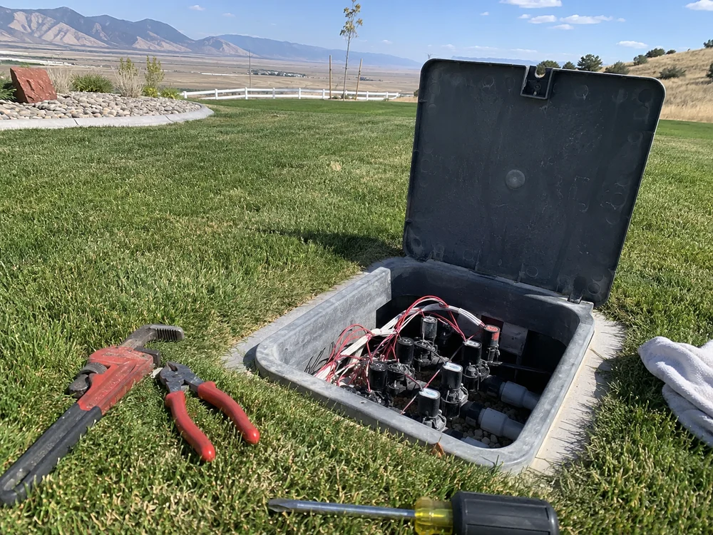Open irrigation valve manifold box with solenoid valves and wiring, tools resting on the grass