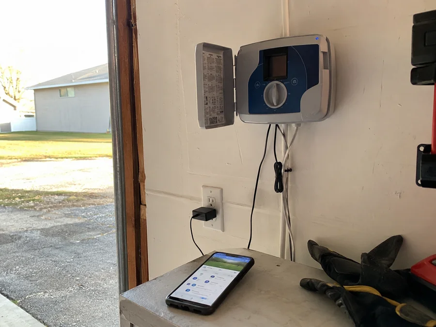 Wall-mounted smart WiFi sprinkler controller in a Utah garage with a phone app open beside it